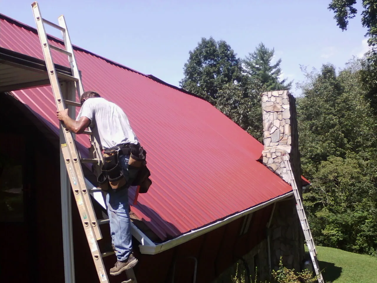 Roofer on a ladder beside a red metal roof and stone chimney — Soffit & Fascia Repair by Roofing Brothers in Winburne